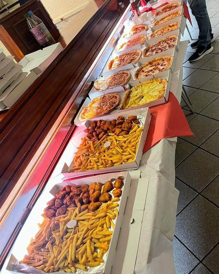 Long table lined with open pizza boxes displaying various pizzas, alongside boxes of fries and fried chicken, set up inside a takeaway or restaurant.